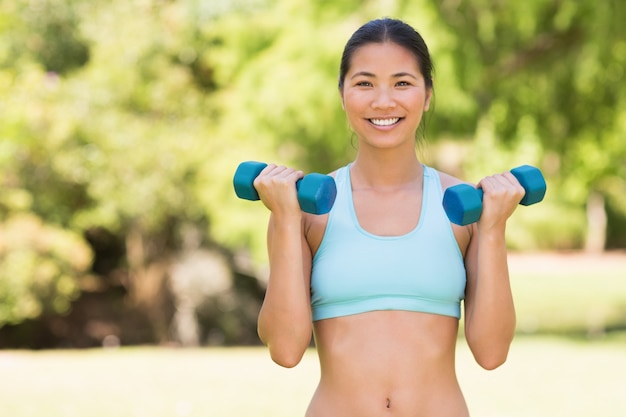 Mujer sonriente sana que ejercita con pesas de gimnasia en parque