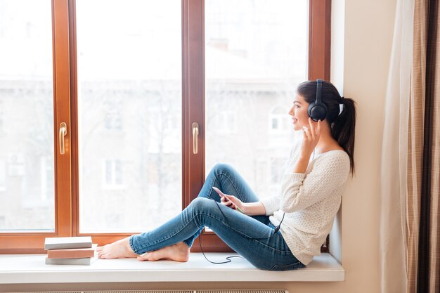 Mujer sonriente sentada en el alféizar de la ventana y escuchando música con auriculares en casa