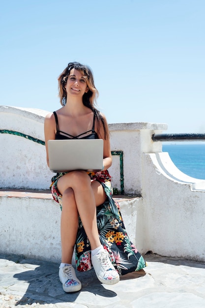 Mujer sonriente sentada en un banco con una computadora cerca del mar