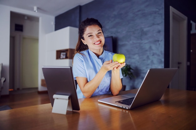 Mujer sonriente sentada en casa rodeada de portátil y tableta y sosteniendo la manzana en las manos