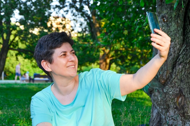 Mujer sonriente sentada en el césped en el parque se toma selfie o se comunica a través de video en un teléfono inteligente