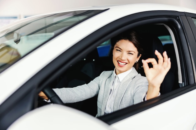 Mujer sonriente sentada en el coche que quiere comprar y sembrando el signo bien.