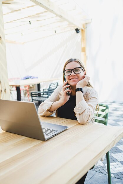 Mujer sonriente sentada en la mesa de un café al aire libre con una computadora portátil hablando por teléfono y una taza de café mujer sonriente con anteojos disfrutando del teletrabajo en un café o estudiando en línea