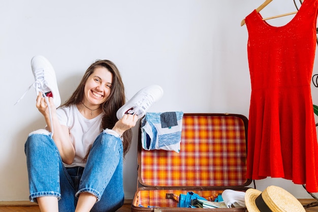 Mujer sonriente sentada en el piso cerca de la maleta con ropa empacada antes del viaje espacio de copia del concepto de viaje