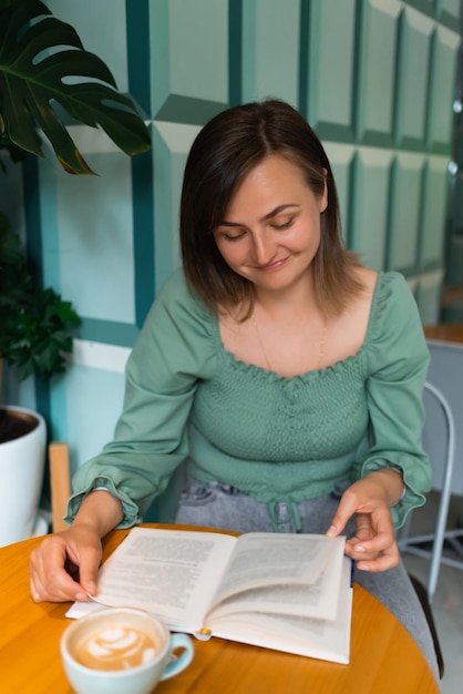 Mujer sonriente sentada en una terraza de verano en la calle con café matutino y leyendo un libro