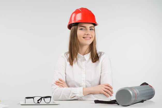 La mujer sonriente se sienta en la mesa con un casco naranja doblado de brazos