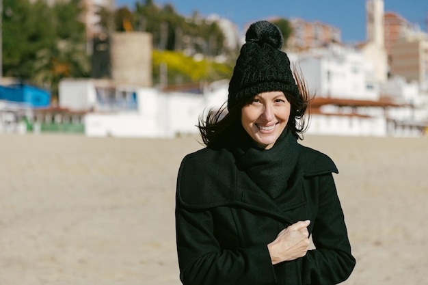 Mujer sonriente con sombrero y abrigo en la playa en un soleado día de invierno