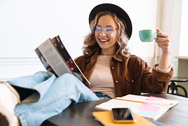 Mujer sonriente con sombrero leyendo una revista con las piernas sobre la mesa mientras bebe café