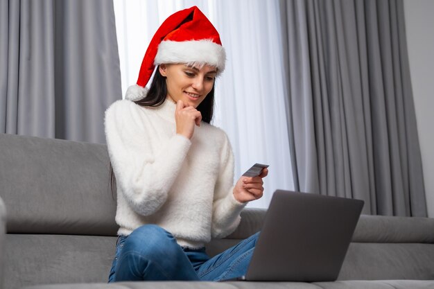 Mujer sonriente con sombrero de Papá Noel sosteniendo una tarjeta de crédito y comprando regalos en línea