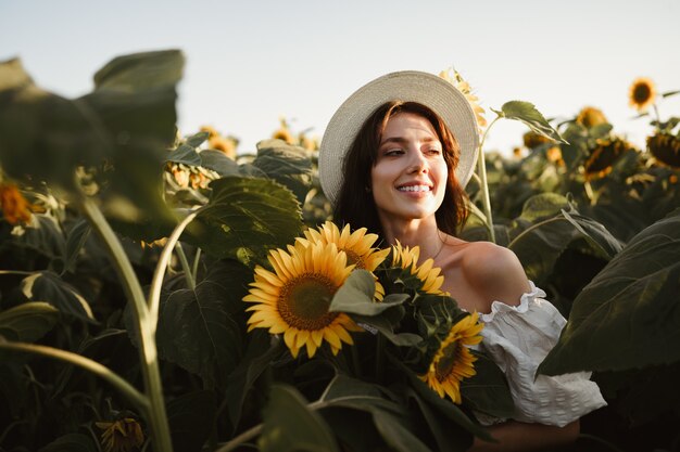 Mujer sonriente con sombrero de pie en un campo de girasoles al atardecer