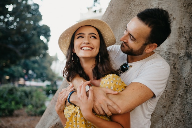 Una mujer sonriente con sombrero y vestido amarillo y su feliz novio con barba se abrazan bajo un viejo Ficus Macrophylla valenciano en España.