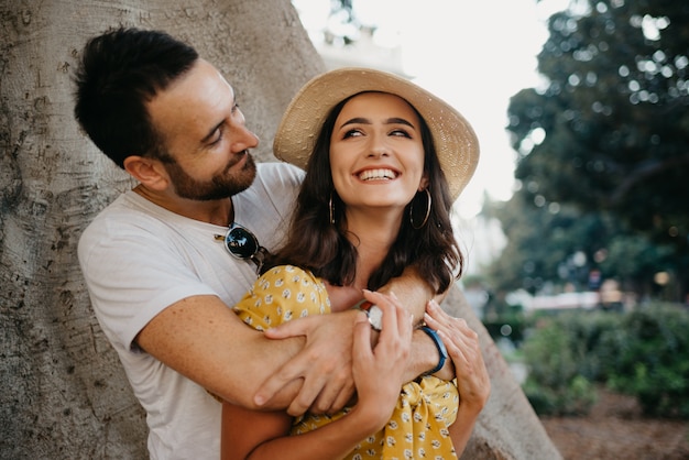 Una mujer sonriente con un sombrero y un vestido amarillo y su feliz novio con barba se abrazan bajo un viejo Ficus Macrophylla valenciano