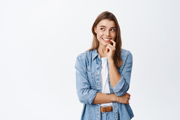 Mujer sonriente soñadora mordiendo el dedo y mirando a un lado, pensando o teniendo una idea interesante, soñando despierto con algo encantador, de pie en blanco