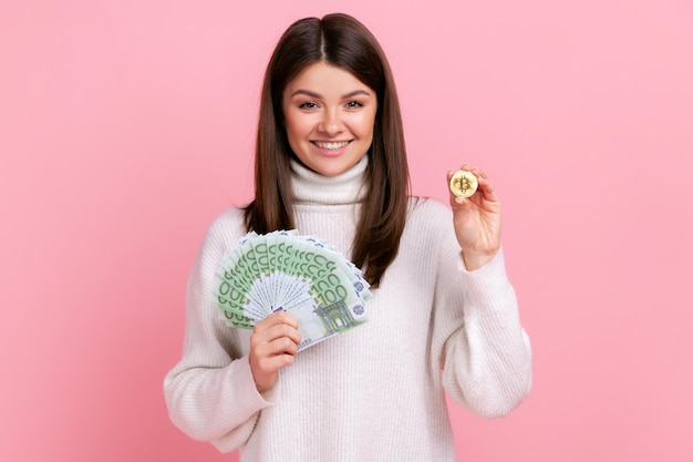 Mujer sonriente sosteniendo bitcoin de oro y una gran suma de dinero, mirando sonriendo a la cámara, gran ganancia, usando un suéter blanco de estilo casual. Disparo de estudio interior aislado sobre fondo rosa.