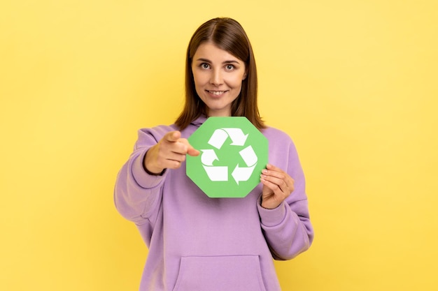 Foto mujer sonriente sosteniendo un cartel de reciclaje verde apuntándote con el dedo, mirando la cámara, pidiendo clasificación de basura y protección del medio ambiente. disparo de estudio interior aislado sobre fondo amarillo.