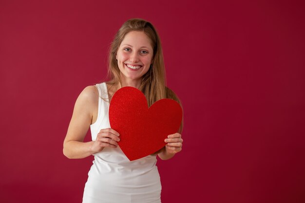 Mujer sonriente sosteniendo corazón papper en manos sobre fondo rojo.