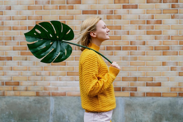 Mujer sonriente sosteniendo una hoja de monstera frente a una pared de ladrillo Concepto de sostenibilidad ecológica