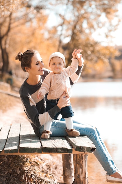 Mujer sonriente sosteniendo a una niña con ropa informal tejida sentada en el parque de otoño Felicidad