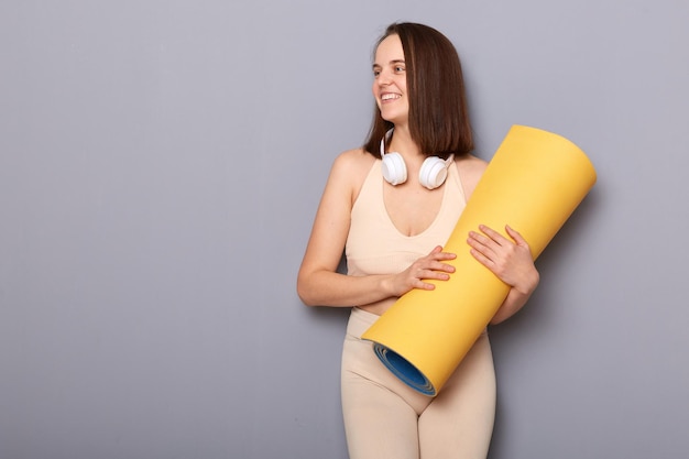 Foto mujer sonriente sosteniendo un tapete de yoga para la meditación mirando hacia otro lado en el espacio de copia de fondo gris para el anuncio de promoción para el gimnasio de ejercicio físico