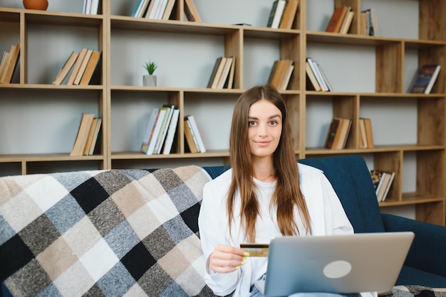 Mujer sonriente sosteniendo una tarjeta de crédito y usando una computadora portátil en el sofá en la sala de estar