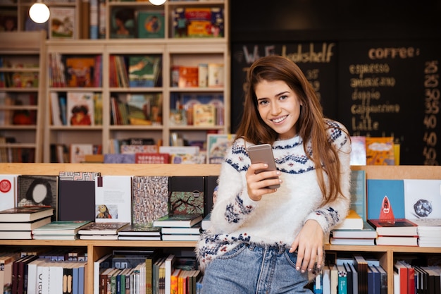 Mujer sonriente sosteniendo un teléfono móvil mientras está sentado en la biblioteca