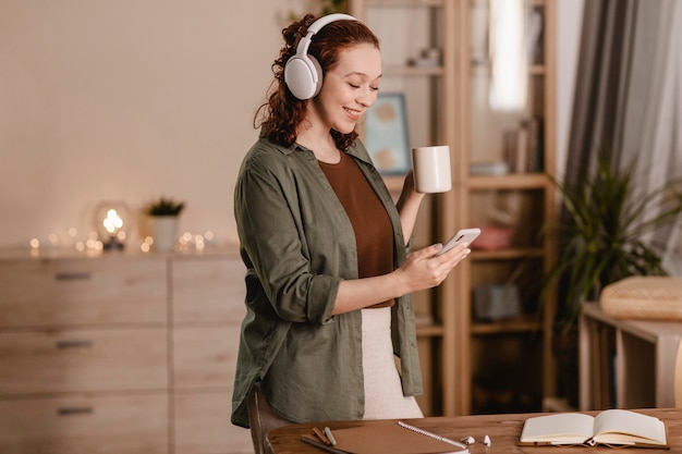 Foto mujer sonriente con su teléfono inteligente y auriculares en casa mientras toma café