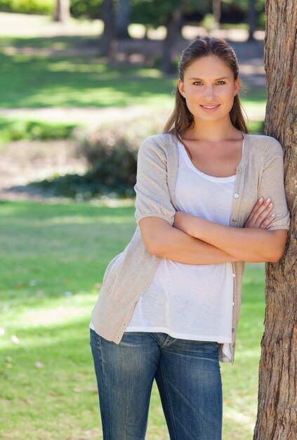 Mujer sonriente con sus brazos cruzados apoyado contra un árbol