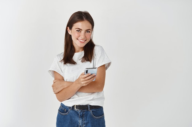 Mujer sonriente con teléfono móvil y mirando al frente feliz, de pie sobre la pared blanca