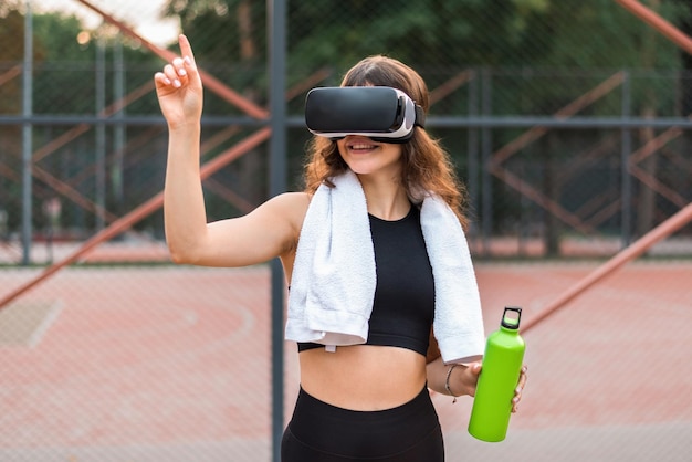 Mujer sonriente con una toalla usando auriculares VR en un chándal sosteniendo una botella de agua en un campo deportivo