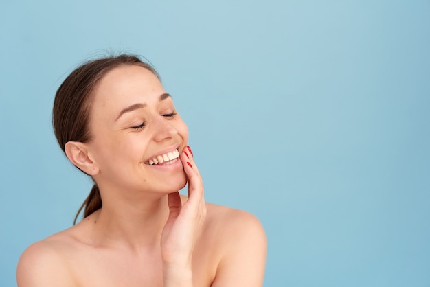 Mujer sonriente tocando su rostro sobre un fondo azul. Concepto de belleza natural, mujer feliz.