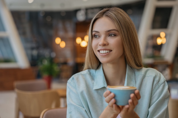 Mujer sonriente tomando café en la cafetería. Taza de explotación femenina feliz con bebida caliente. Concepto de pausa para el café