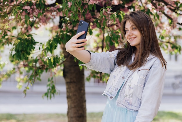 Mujer sonriente tomando fotos de autorretrato selfie en modelo de smartphone posando en el parque de árboles de sakura