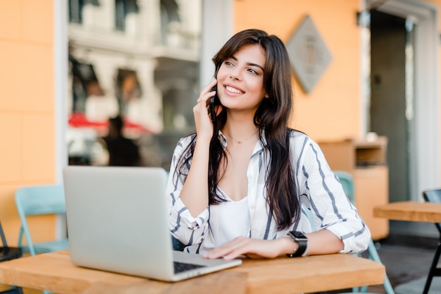 La mujer sonriente usa la computadora portátil que se sienta cerca de café