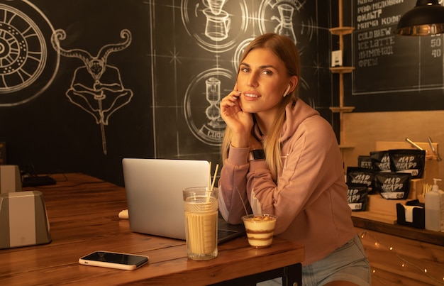 Mujer sonriente usa computadora portátil, se sienta en la cafetería con una taza de capuchino y postre