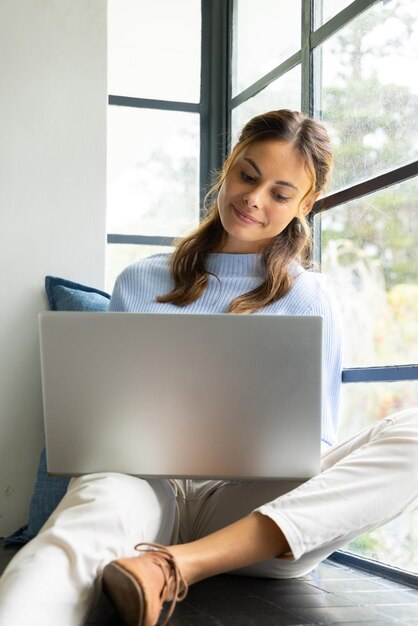 Mujer sonriente usando una computadora portátil junto a la ventana disfrutando de una acogedora atmósfera doméstica