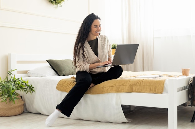 Mujer sonriente usando una laptop sentada en la cama en casa