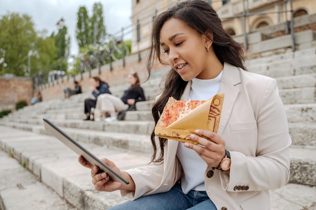 Mujer sonriente usando una tableta digital mirando la pantalla, sosteniendo una rebanada de pizza sabrosa al aire libre