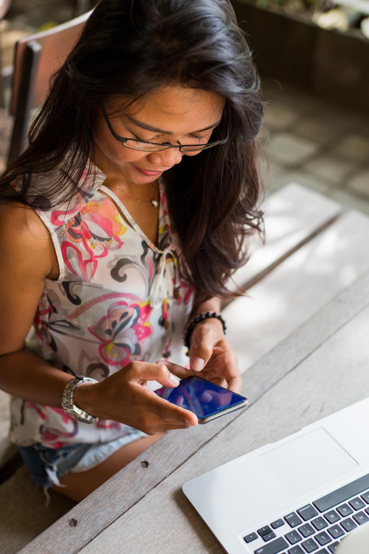 Foto mujer sonriente usando el teléfono móvil mientras está sentada junto a la computadora portátil en la mesa