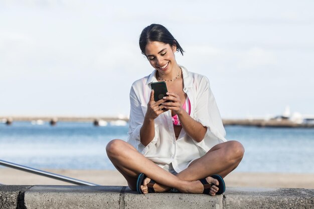 Foto mujer sonriente usando el teléfono móvil mientras está sentada contra la bahía