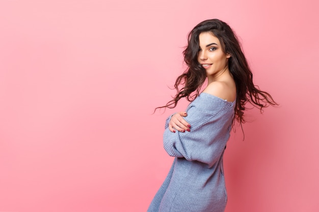 Mujer sonriente vestida con suéter de punto azul mirando por encima del hombro mientras posando sobre fondo rosa