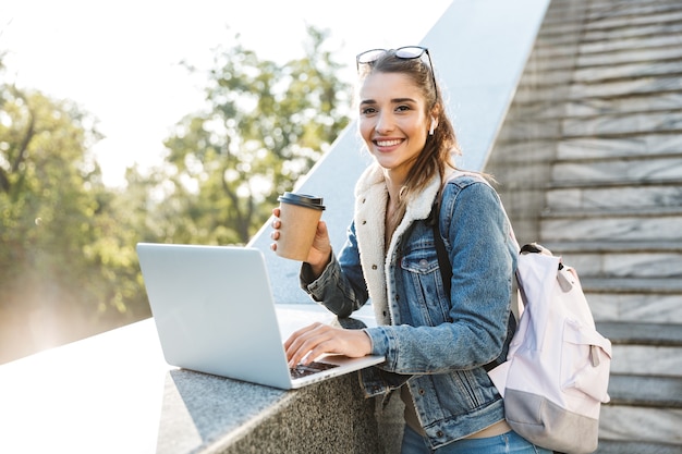 Mujer sonriente vistiendo chaqueta de pie en las escaleras al aire libre, utilizando equipo portátil, sosteniendo una taza de café