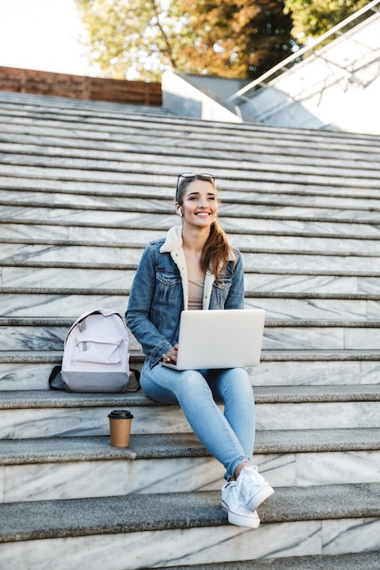 Mujer sonriente vistiendo chaqueta sentada en las escaleras al aire libre, utilizando equipo portátil