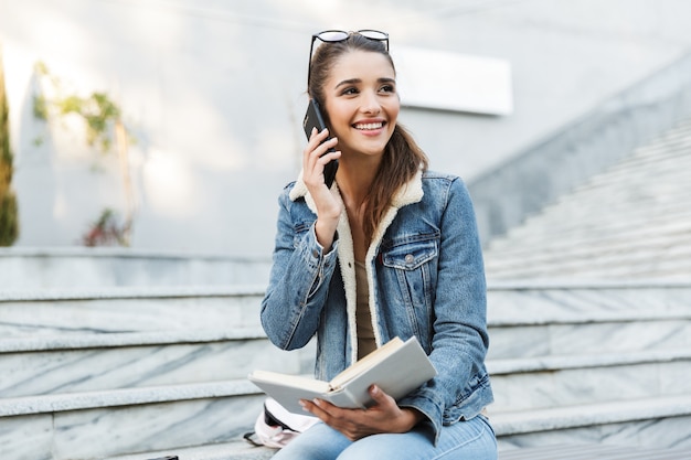 Mujer sonriente vistiendo chaqueta sentado en un banco al aire libre, libro de lectura, hablando por teléfono móvil