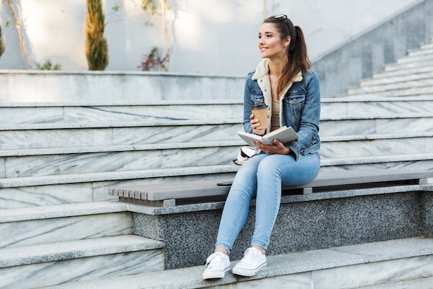 Mujer sonriente vistiendo chaqueta sentado en un banco al aire libre, libro de lectura, sosteniendo una taza de café para llevar