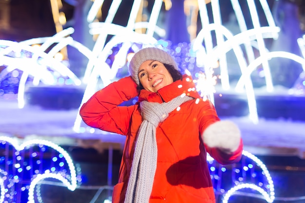 Mujer sonriente vistiendo ropa de punto de invierno sosteniendo bengala al aire libre sobre fondo de nieve