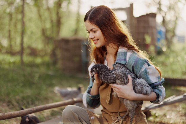 Foto una mujer con una sonrisa cuida de un pollo saludable y sostiene un pollo en sus manos mientras trabaja en una granja en la naturaleza alimentando con comida orgánica a las aves bajo el sol