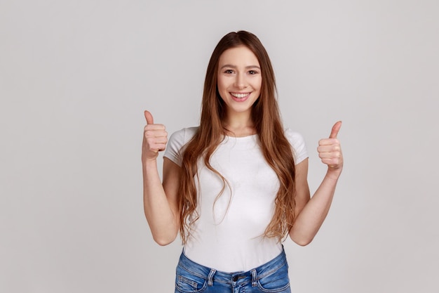 Mujer con sonrisa dentuda, mostrando los pulgares hacia arriba, signo de aprobación, satisfecha con el servicio, buena retroalimentación, expresando emociones positivas, usando una camiseta blanca. Disparo de estudio interior aislado sobre fondo gris.