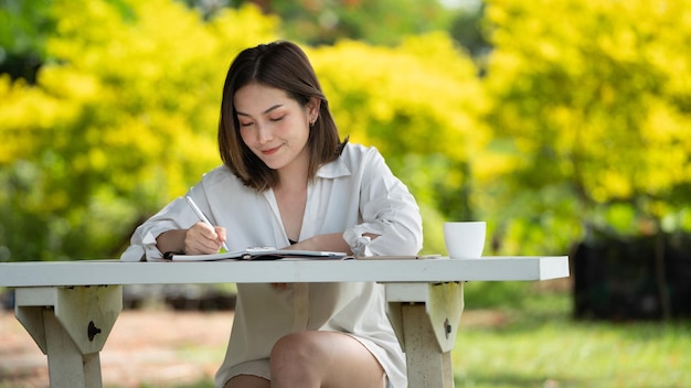 Mujer de sonrisa pensativa en el parque usando un cuaderno y escribiendo Retrato de una joven mujer de negocios encantadora revisando el trabajo de negocios en línea en su teléfono inteligente al aire libre en el parque en un fondo verde suave