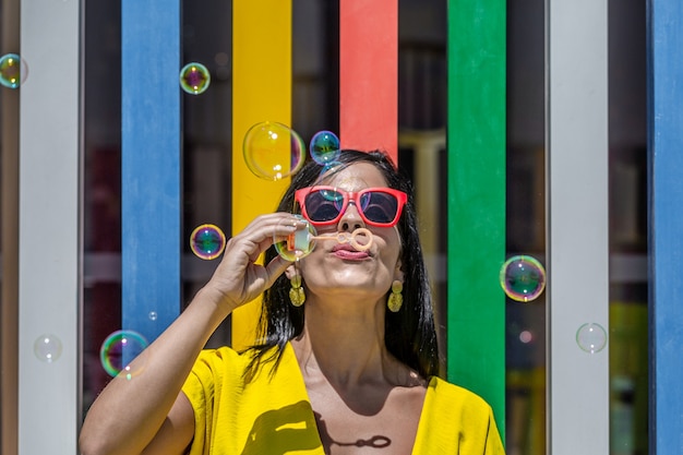 Foto mujer soplando burbujas de jabón hacia arriba en un vestido amarillo y gafas de sol