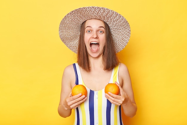 Mujer sorprendida alegre con traje de baño a rayas y sombrero aislado en un fondo amarillo sosteniendo naranjas gritando de emoción